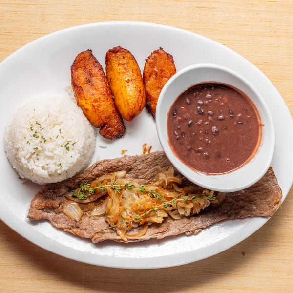Cuban steak with caramelized onions, white rice, black beans, and fried plantains on a white plate.