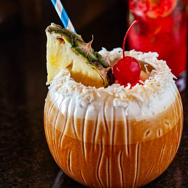 A tropical cocktail in a coconut-shaped glass, garnished with pineapple, cherry, and shredded coconut.