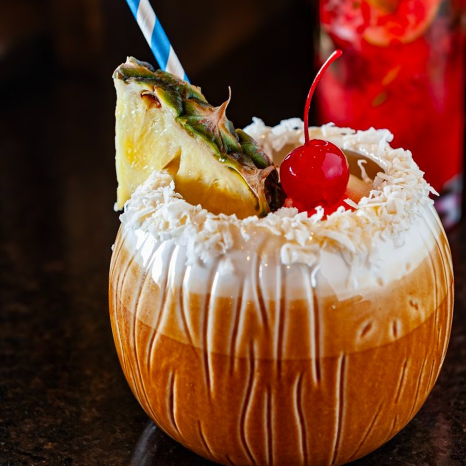 A tropical cocktail in a coconut-shaped glass, garnished with pineapple, cherry, and shredded coconut.