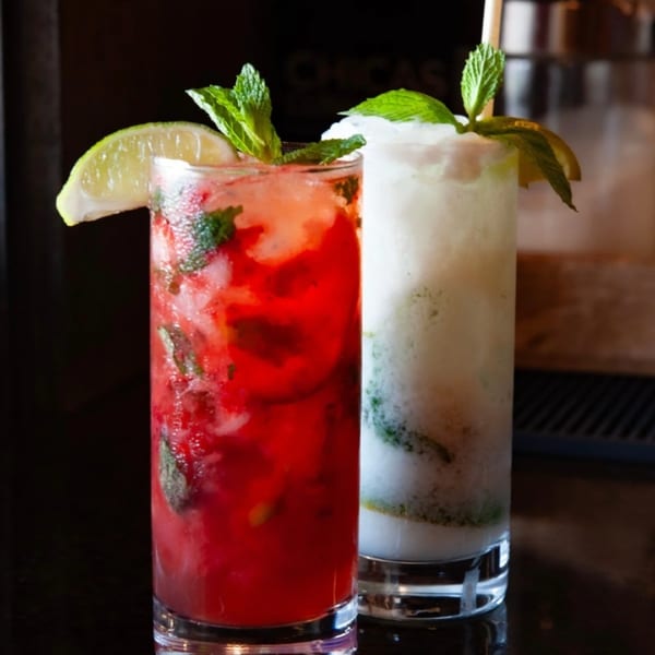 Two mojitos, one red and one white, garnished with lime and mint, on a dark bar.
