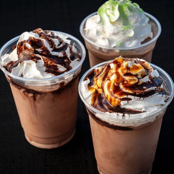Three Cuban cafe milkshakes with whipped cream, chocolate, caramel, and green drizzle on a dark surface.