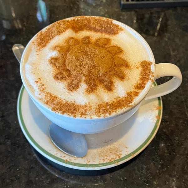 A white cup of coffee with a cinnamon sun design on top, on a saucer with a spoon.